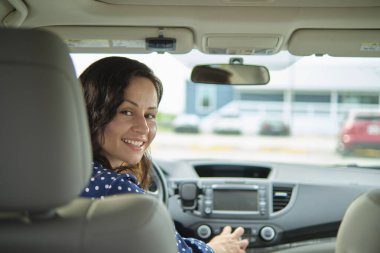 Direksiyon başında oturup araba sürerken omzunun üstünden bakan sıradan giysili çekici genç bir kadının portresi..