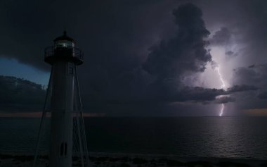 Deniz kıyısındaki uzun deniz feneri fırtınalı bir gecede ticari gemilerin seyrüsefer için yanıp sönen ışık. Okyanus üzerinde şimşekler çakan fırtına gemiler için tehlike arz ediyor..