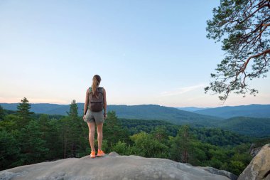 Sportive woman walking alone on hillside trail. Female hiker enjoying view of evening nature from rocky cliff on wilderness path. Active lifestyle concept.