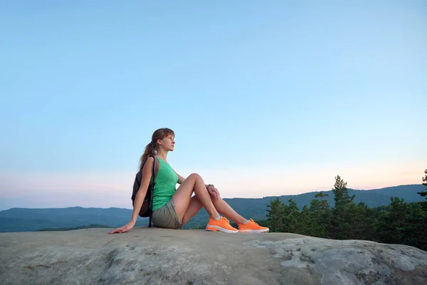Hiker girl resting on rocky mountain top enjoying morning nature during her travel on wilderness trail. Lonely female traveler traversing high hilltop route. Healthy lifestyle concept.
