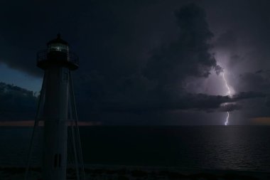 Deniz kıyısındaki uzun deniz feneri fırtınalı bir gecede ticari gemilerin seyrüsefer için yanıp sönen ışık. Okyanus üzerinde şimşekler çakan fırtına gemiler için tehlike arz ediyor..