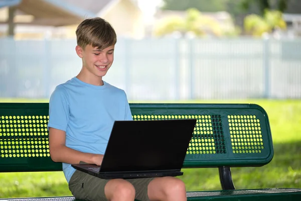 Schoolboy studying on laptop computer resting on park bench outdoors on summer day. Remote education during quarantine concept.