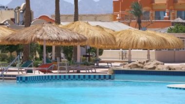 Boş güverte sandalyeleri, tropik tatil köylerinde havuz kenarında saman gölgesi şemsiyelerinin altında. Yaz tatilleri ve kaçış konsepti.