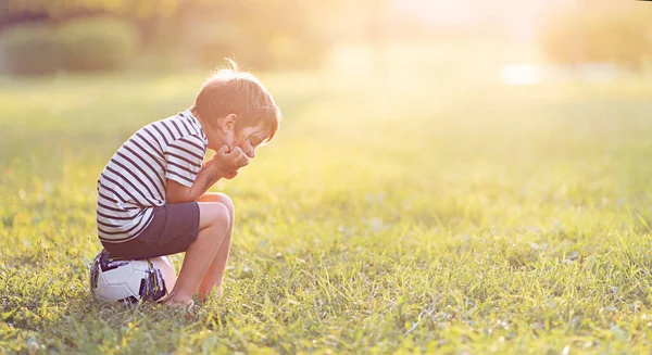 Mały chłopiec siedzi i jest smutny na trawiastym polu z promykiem słońca. — Zdjęcie stockowe