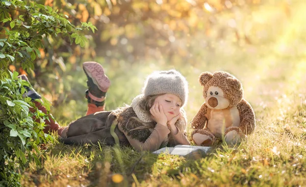Little girl reading a book in a forest clearing with her teddy bear in autumn, fairy tale picture — Stockfoto
