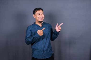 Gri arka planda izole edilmiş genç Asyalı bir adam bir ürün sunmak için kenarı işaret ediyor.