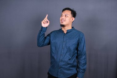Gri arka planda izole edilmiş genç Asyalı bir adam bir ürün sunmak için kenarı işaret ediyor.