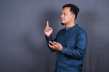 Gri arka planda izole edilmiş genç Asyalı bir adam bir ürün sunmak için kenarı işaret ediyor.