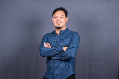 Mavi tişörtlü gülümseyen Asyalı adam elleri katlanmış gri arka planda tek başına duruyor. Mühendislik işleri kavramı