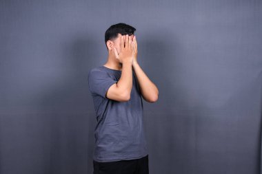 Depresyondaki Asyalı genç adam ellerini yüzünü kapatarak stresli zaman geçiriyor. Gri arkaplanda izole. 