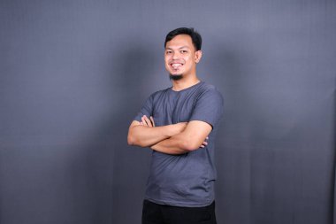 Gri tişörtlü gülümseyen yakışıklı kollarını kavuşturmuş gri arka planda duruyor.