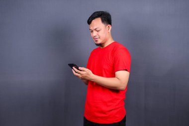 Kırmızı tişört giyen neşeli bir Asyalının portresi. Gri arka planda izole bir şekilde duruyor. Cep telefonu kullanıyor.