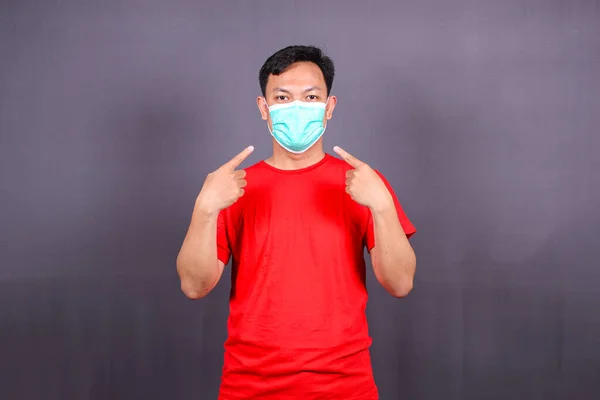 Asian young man pointing at face mask, asking to use measures against covid-19,  isolated on gray background