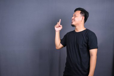 Siyah tişörtlü mutlu bir Asyalı 'nın portresi gri arka planda izole edilmiş fotokopi alanını işaret ediyor.