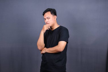 Genç Asyalı adam çenesini tutuyor ve kameraya dargın bakıyor. Öfkeli uyanışlar, kötü ruh halinde, gri arka planda izole edilmiş.