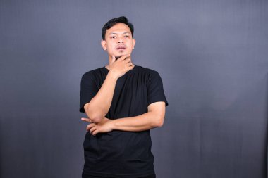 Genç Asyalı adam çenesini tutuyor ve kameraya dargın bakıyor. Öfkeli uyanışlar, kötü ruh halinde, gri arka planda izole edilmiş.