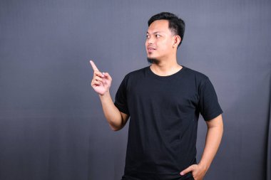 Siyah tişörtlü mutlu bir Asyalı 'nın portresi gri arka planda izole edilmiş fotokopi alanını işaret ediyor.