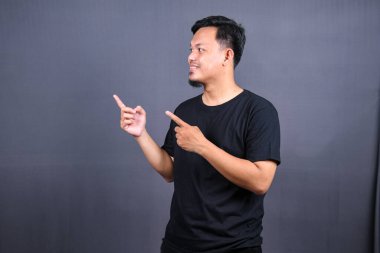 Siyah tişörtlü mutlu bir Asyalı 'nın portresi gri arka planda izole edilmiş fotokopi alanını işaret ediyor.