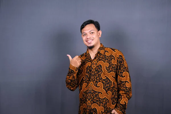 一个穿着蜡染印度尼西亚的快乐年轻人的画像 手指指向灰色背景上孤立的复制空间 — 图库照片