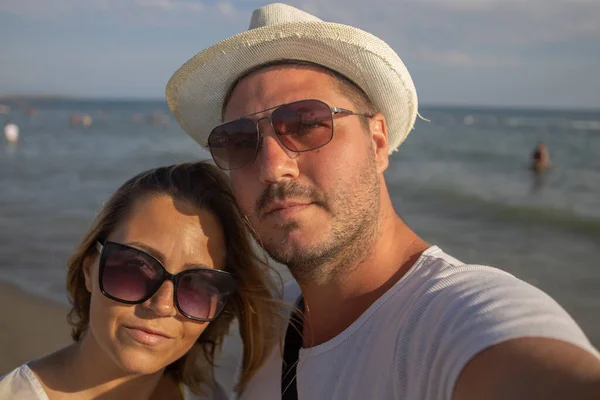 Genç evli bir çift sahilde poz veriyor ve fotoğraf çekiyor. Adam ve kadın güneş gözlüğü takıyor ve adamın şapkası var.
