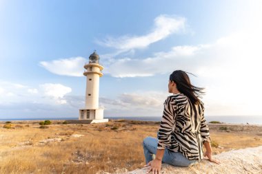 Young woman sits looking at a lighthouse. She is on a trip and visits the place to watch the sunset. She is in Barbaria, Formentera, Balearic Islands. clipart