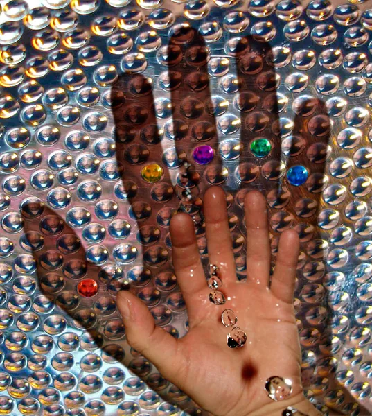 Multiple reflections of a face and a hand levitating drops of water frozen in time with the silhouette behind. At each end of each finger is a primary color face.