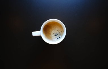 Masanın üzerinde köpüklü kahve olan beyaz bir fincan. Yukarıdan bak. Siyah masa. Köpüklü Americano.