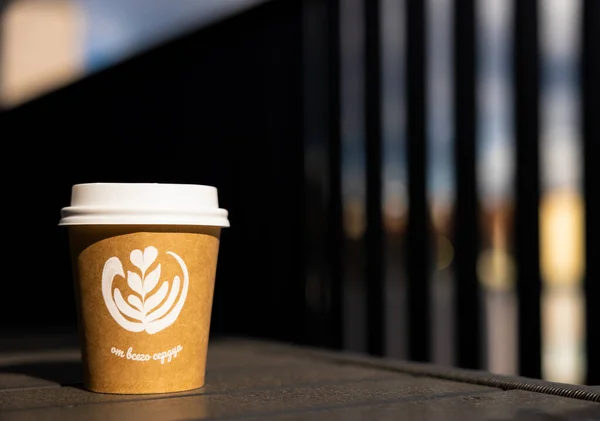 A paper cup of coffee with a white lid stands on a table in the sun. Table on the open veranda of the cafe. the inscription is from the bottom of my heart. Translation - from the bottom of my heart.