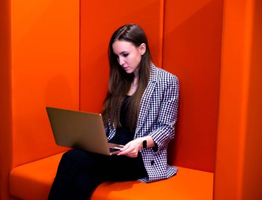 Ciddi bir kadın dizüstü bilgisayar kullanıyor, internetten e-posta haberlerini kontrol ediyor, turuncu koltukta oturuyor, bilgisayar başında çalışıyor, blog yazıyor ya da webinar izliyor, ders çalışıyor. Yüksek kalite fotoğraf