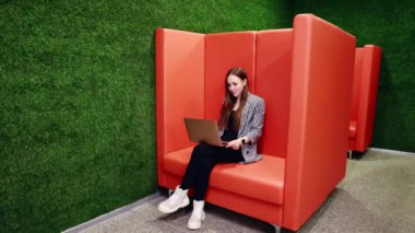 Kız bir dizüstü bilgisayarda uzak bir toplantı düzenliyor. Ofisteki turuncu bir kanepede oturmak. Yüksek kaliteli FullHD görüntüler