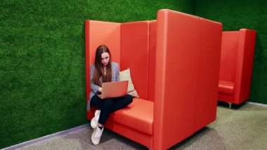 Kız, dinlenmek için esnediği işi yaptıktan sonra dizüstü bilgisayarla çalışıyor. Ofisteki turuncu bir kanepede oturmak. Yüksek kaliteli FullHD görüntüler