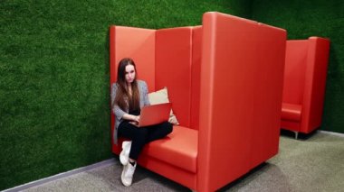 Kız bir dizüstü bilgisayarla çalışıyor, yandan dalgın dalgın bakıyor. Ofisteki turuncu bir kanepede oturmak. Yüksek kaliteli FullHD görüntüler