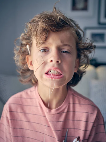 Cute boy with plaster on cheek and thread on tooth looking at camera while standing in light room at home