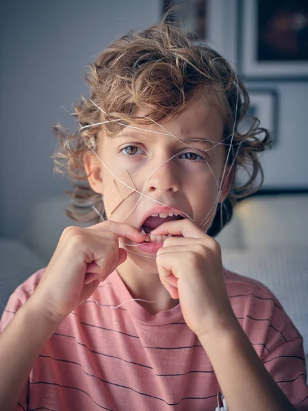 Brave boy in casual wear with thread on face looking away while touching loose tooth in light living room at home