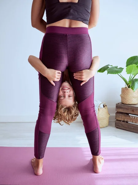 Crop woman in sportswear holding preteen child upside down while practicing acro yoga in lounge room at home
