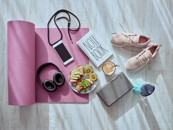 Top view of assorted fruits and coffee placed near smartphone and headphones on yoga mat with notebook and sneakers in light room