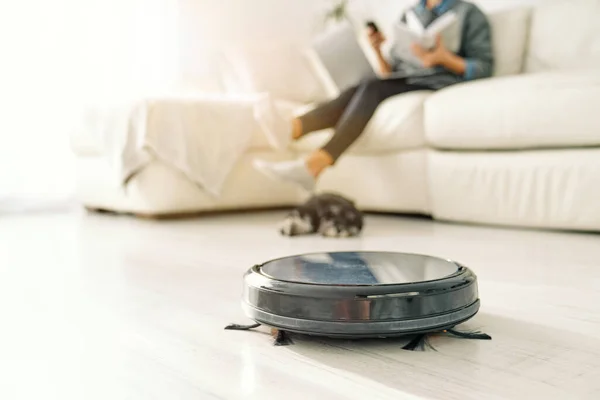 Black robotic vacuum cleaner cleaning floor against sleeping dog and person with book sitting on sofa in living room