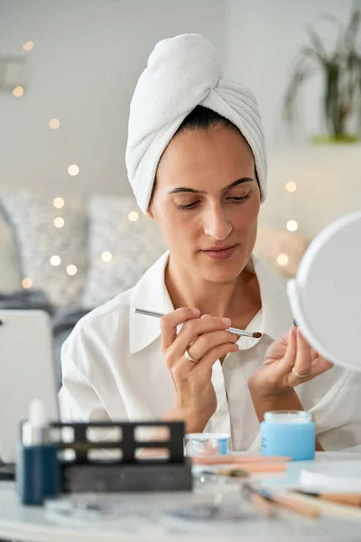 Focused adult female blogger with towel on head doing makeup with cosmetic brush in front of mirror while shooting beauty blog on smartphone at home in morning