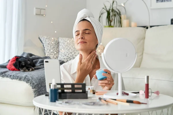 Relaxing female beauty blogger applying cosmetic cream on neck while recording video for vlog via smartphone