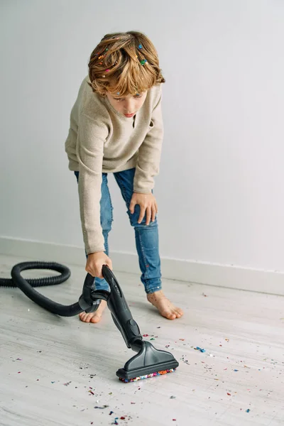 Full body of barefoot boy tidying floor covered with scattered confetti with vacuum cleaner after party