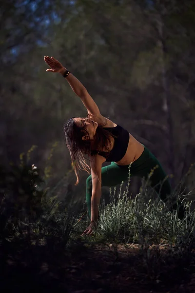 Sportive Hispanic female in sports top and leggings extending arm to sky while performing Parshvakonasana posture during yoga practice in nature