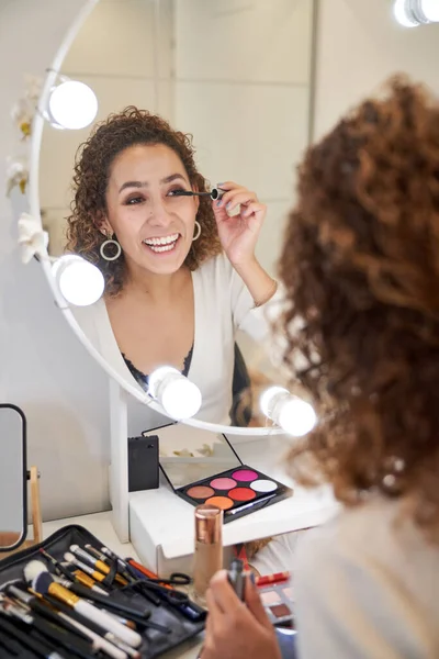 Smiling female beautician putting mascara on eyelashes while doing eye makeup looking in mirror with illuminated lamps