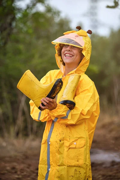 Smiling child with yellow gumboots and in raincoat looking away while standing on muddy pathway in woods on rainy day