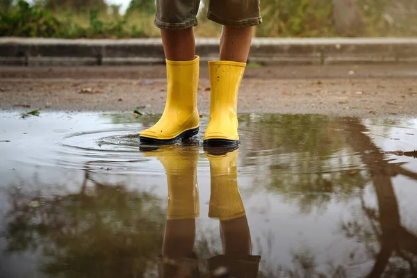 Crop anonymous child wearing yellow rubber boots standing in puddle on asphalt road near forest