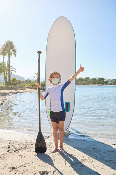 Full length of preteen kid in mask with wet hair standing on sandy beach near SUP board and holding paddle while raising hand and showing like gesture