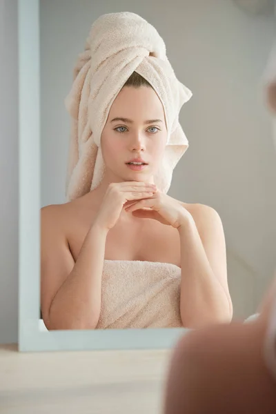 Young female model with towel wrapped around head standing in front of mirror and touching chin while admiring reflection in morning