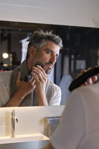 Serious man with towel looking at reflection in mirror and trimming beard in modern bathroom at home