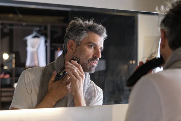 Focused man with gray beard doing morning routine and shaving using professional trimmer while looking at mirror in modern bathroom at home