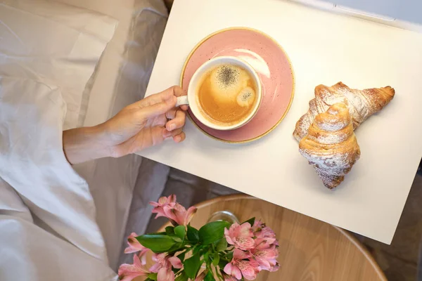 Top view of crop woman lying on soft white bed covered with duvet and reaching out coffee mug and croissants placed on white table in bedroom at home