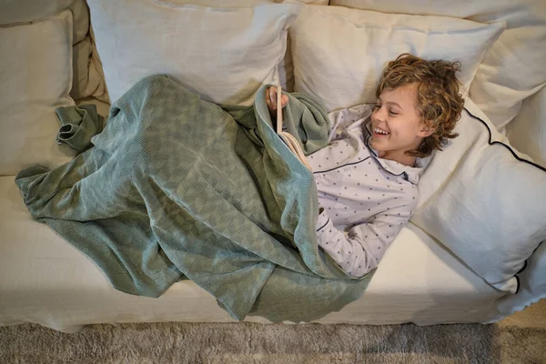 From above full body of cheerful boy in pajama reading book while resting on comfortable couch in light living room